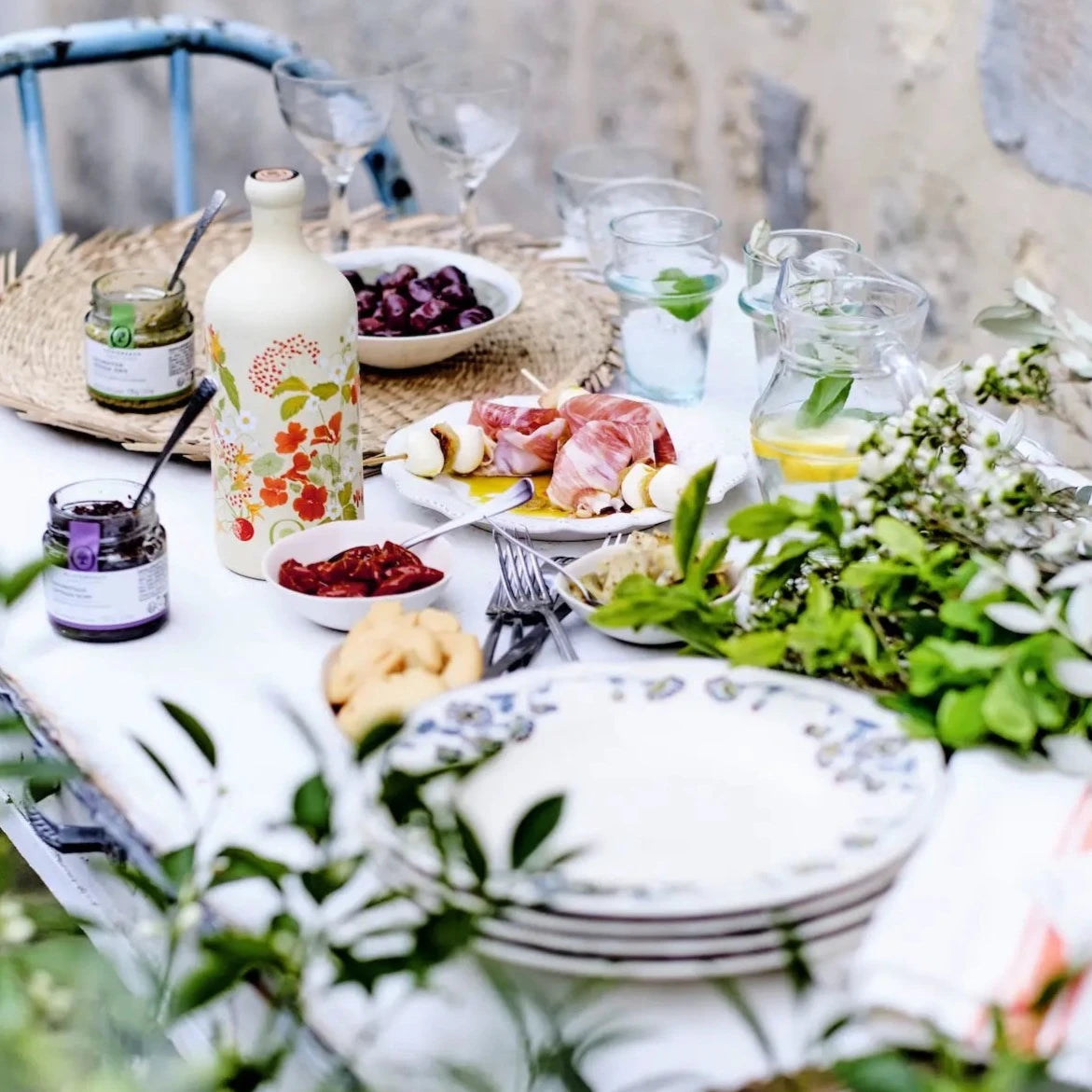 olivenöl flasche limited edition sommer aus italien mit blumen print darauf auf einem Tisch mit antipasti, Oliven und Schinken